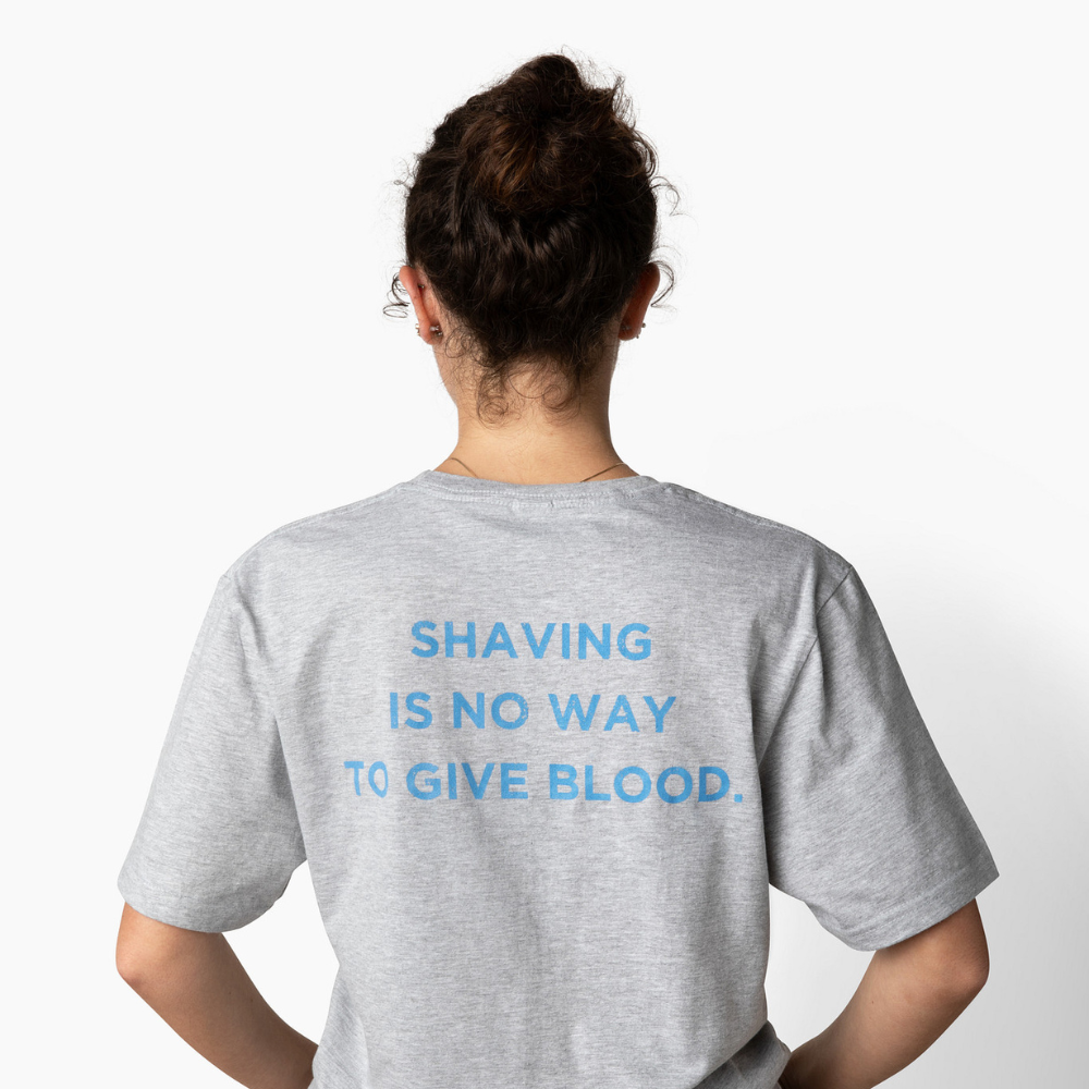 Woman wearing a gray t-shirt with text on the back that says "Shaving is no way to give blood" against a white background