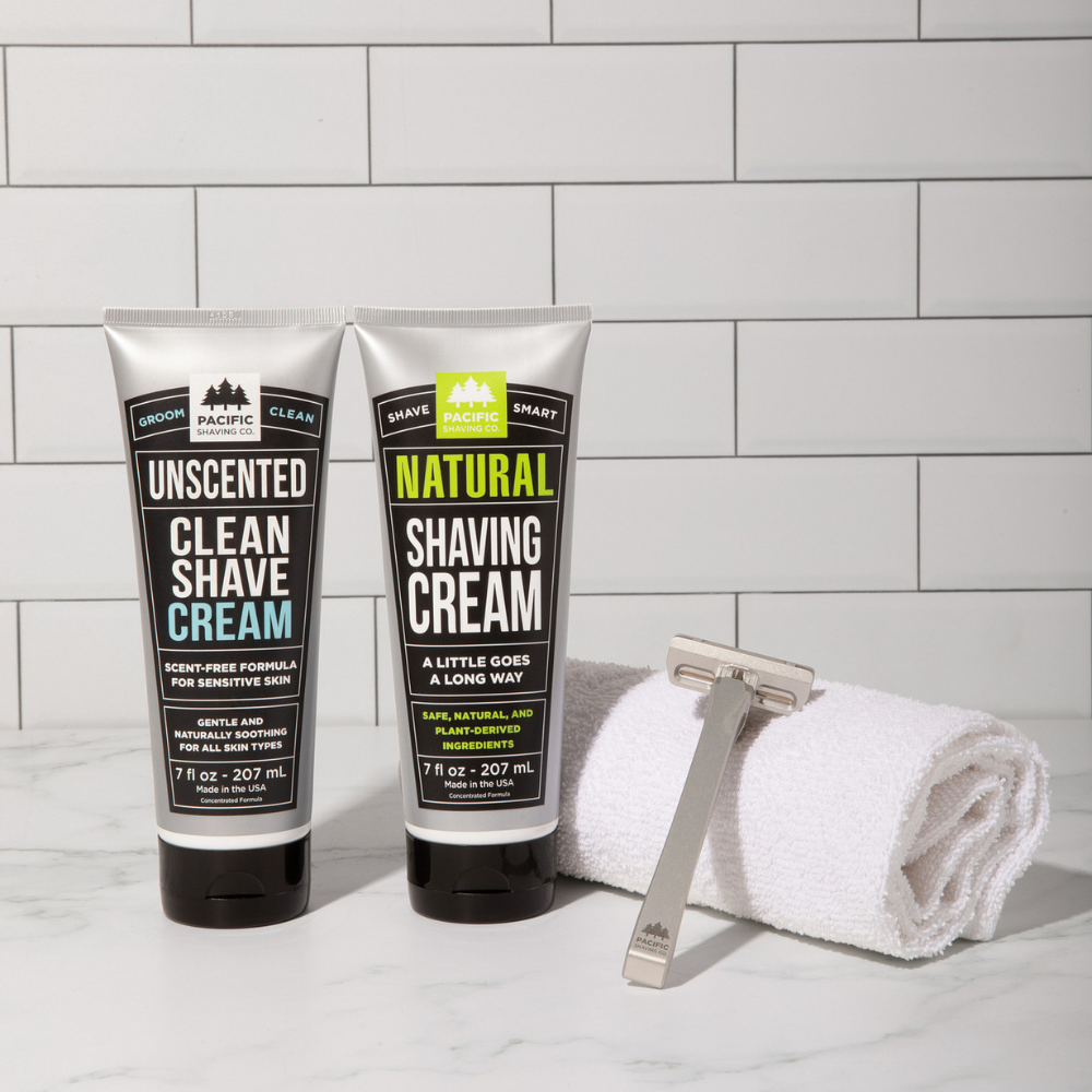 Two tubes of Shave Cream with a razor and towel on a tiled bathroom counter.
