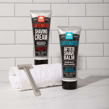 Two tubes of Caffeinated Shaving Cream and After Shave Balm with a razor and towel on a tiled bathroom counter.