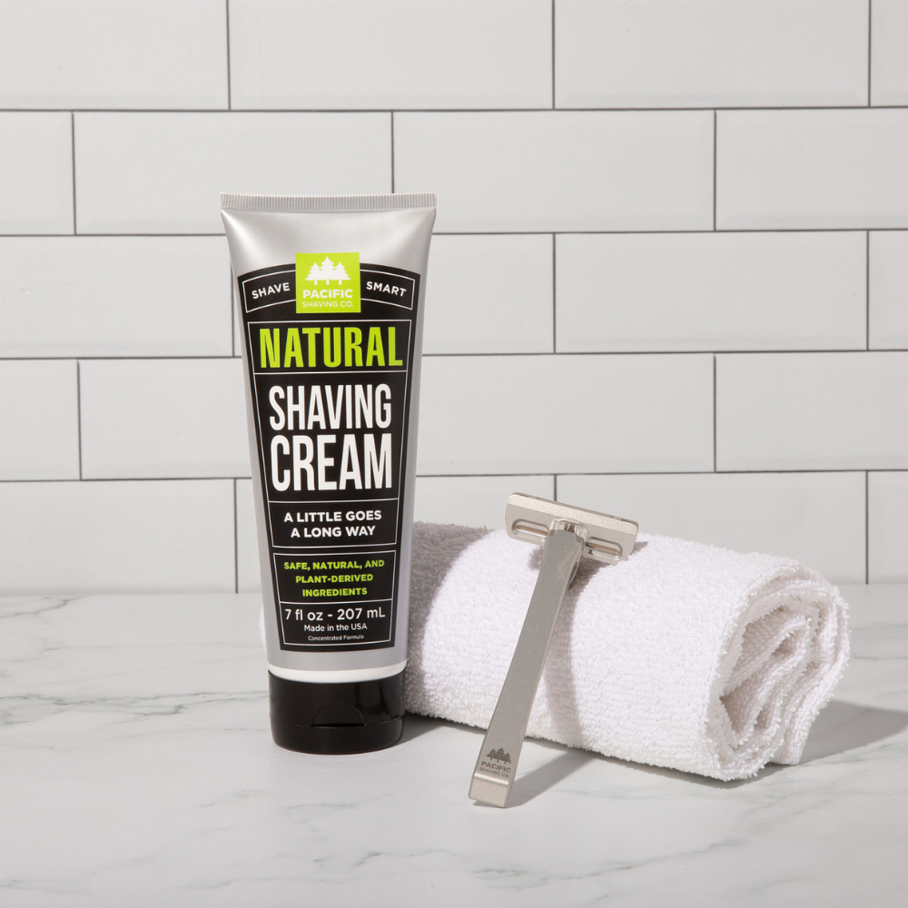 Tube of natural shaving cream with a razor and towel on a tiled bathroom counter.