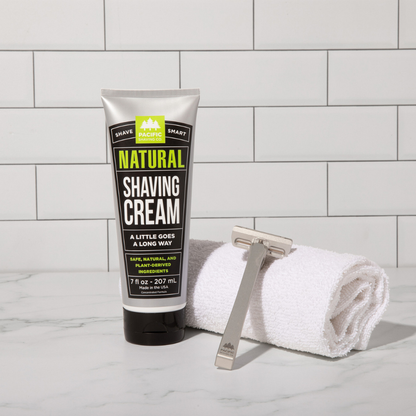 Tube of natural shaving cream with a razor and towel on a tiled bathroom counter.