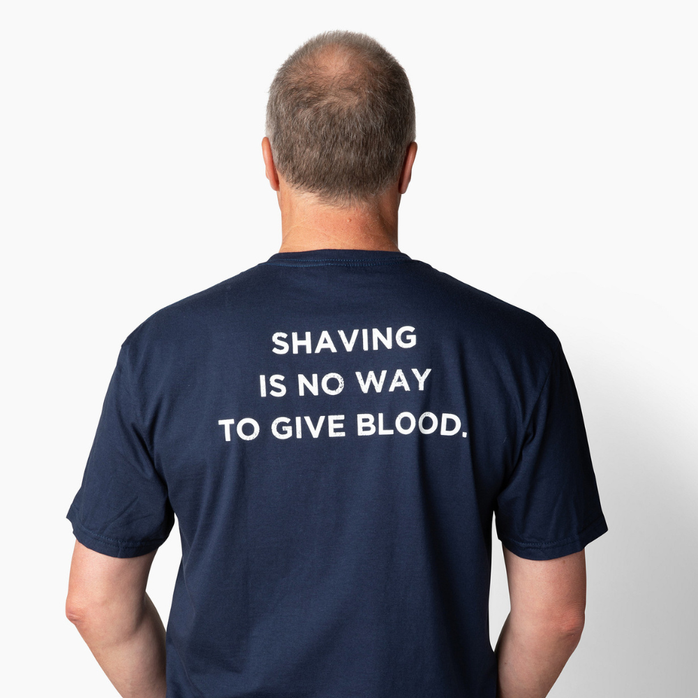 Man wearing a navy blue t-shirt with text on the back that says "Shaving is no way to give blood" against a white background
